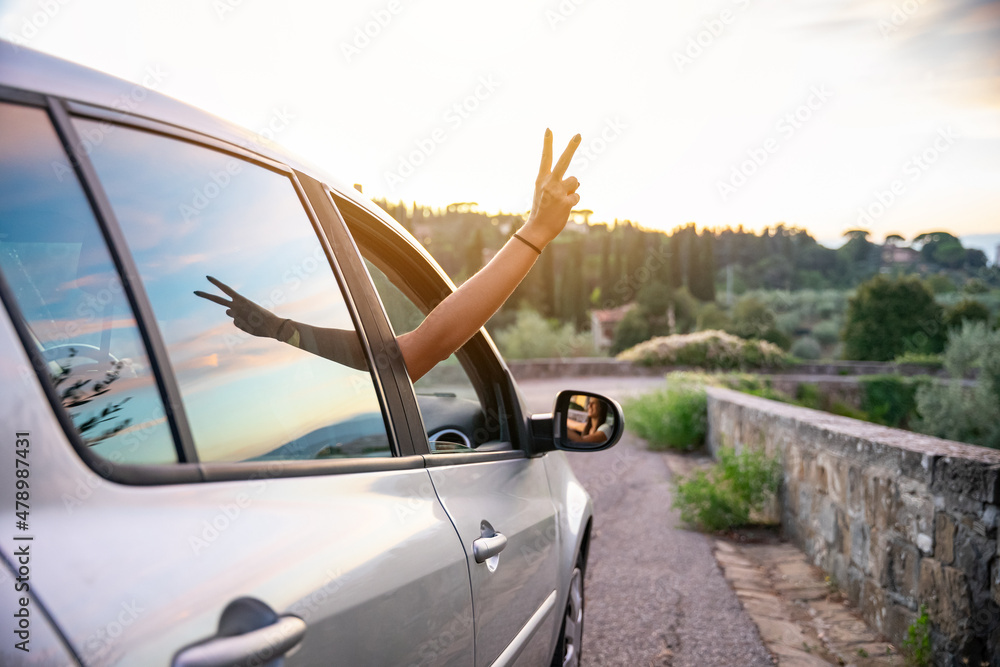 Two friends on holiday in the summer at sunset - Millennial show fingers up from the car window - Relaxed woman during a free trip in the nature - People happy for adventures and carefree leisure