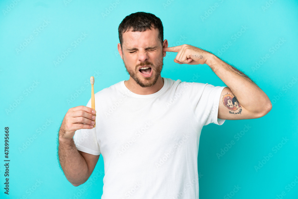 Young Brazilian man brushing teeth isolated on blue background frustrated and covering ears Young Brazilian man brushing teeth isolated on blue background frustrated and covering ears