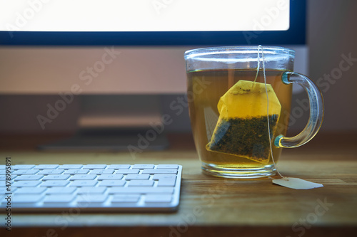 Work in the evening with a cup of tea with a visible tea bag. Keyboard and monitor out of focus. People work until late in the evening in the office or in the home office. Copy space background.