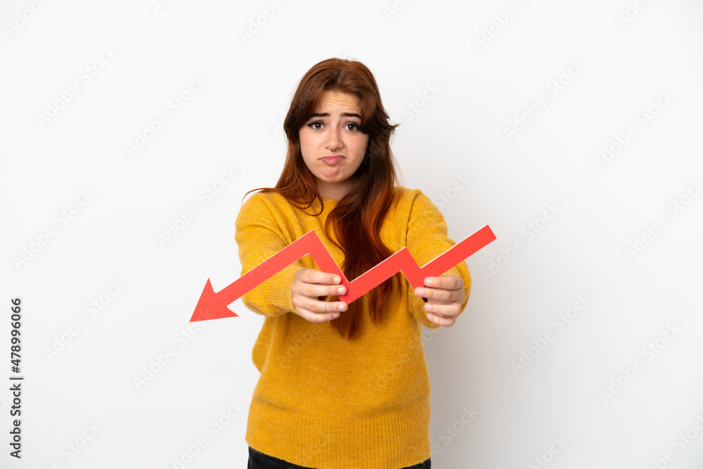 Young redhead woman isolated on white background holding a downward arrow and with sad expression