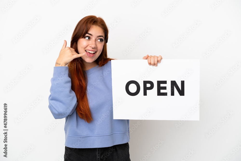 Young redhead woman isolated on white background holding a placard with text OPEN and doing phone gesture Young redhead woman isolated on white background holding a placard with text OPEN and doing phone gesture