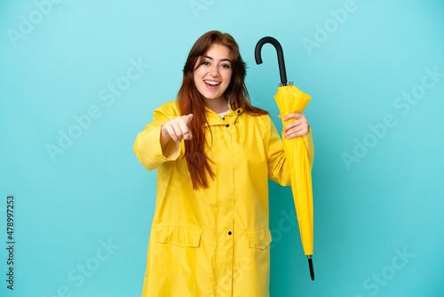 Redhead woman holding an umbrella isolated on blue background surprised and pointing front
