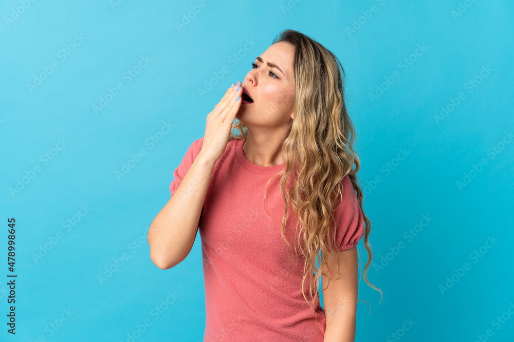 Young Brazilian woman isolated on blue background yawning and covering wide open mouth with hand Young Brazilian woman isolated on blue background yawning and covering wide open mouth with hand