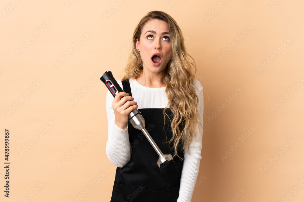 Young brazilian woman using hand blender isolated on beige background looking up and with surprised expression Young brazilian woman using hand blender isolated on beige background looking up and with surprised expression