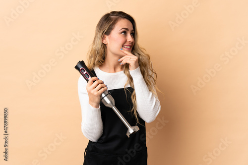 Young brazilian woman using hand blender isolated on beige background looking up while smiling