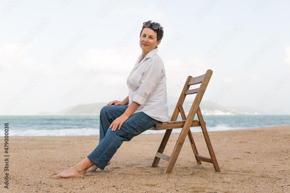 Happy retired woman wearing shawl sitting on chair relaxed on sand at the beach. Senior caucasian woman
