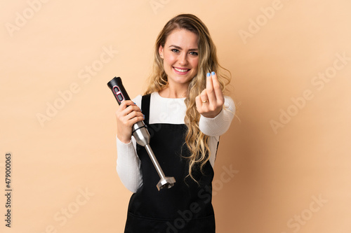 Young brazilian woman using hand blender isolated on beige background making money gesture