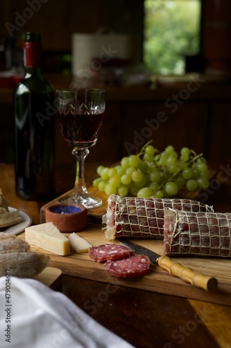 A sausage roll of wild boar salami sliced on a wooden board and being served with cheese, wine and grapes on a wooden table.