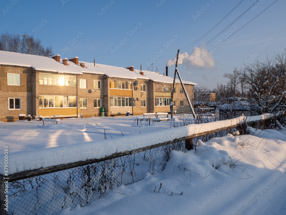 Winter evening in a snowy village. The rays of the setting sun illuminate houses, fence, vegetable gardens and greenhouse.