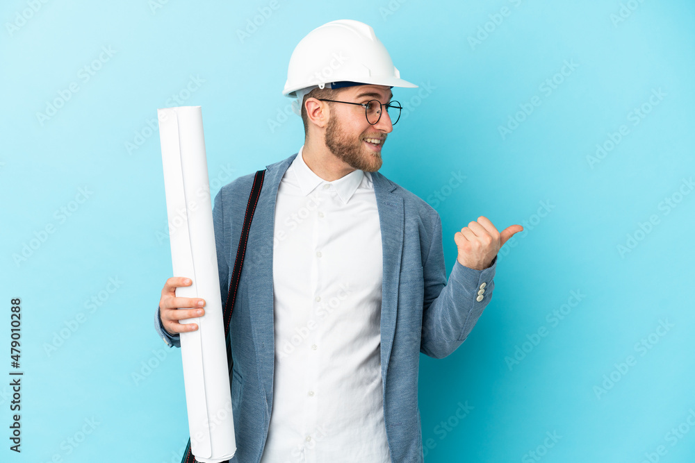 Young architect man with helmet and holding blueprints over isolated background pointing to the side to present a product