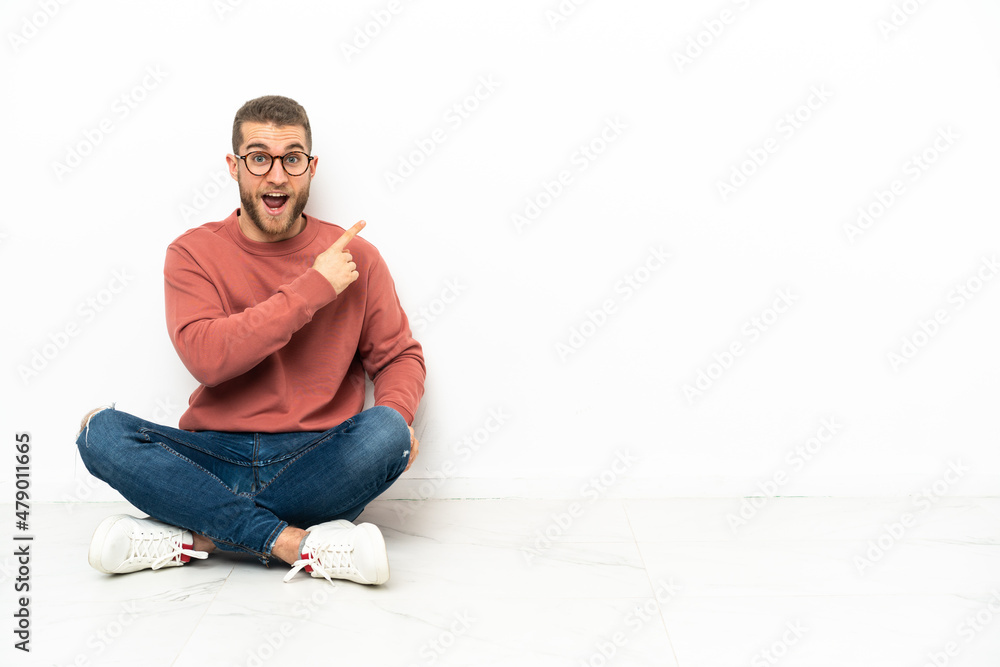 Young handsome man sitting on the floor surprised and pointing side Young handsome man sitting on the floor surprised and pointing side