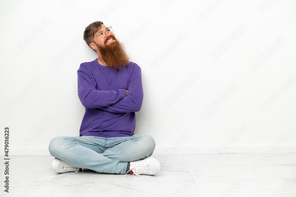 Young caucasian reddish man sitting on the floor isolated on white background looking up while smiling