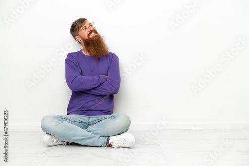 Young caucasian reddish man sitting on the floor isolated on white background looking up while smiling © luismolinero