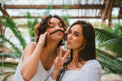 Two funny teenager girls looking at camera and smiling. One of them is playing and making mustache of other girl's hair. photo