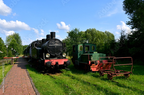 A view of a well maintained public park full of shrubs, trees, and grass, with a train station located nearby and an old traditional steam locomotive waiting for departure seen on a sunny summer day