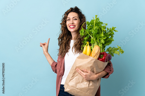 Young woman holding a grocery shopping bag isolated on blue background pointing back