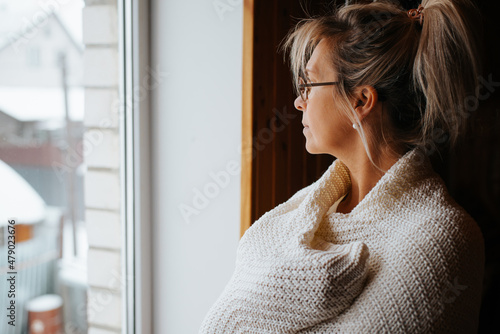 Side view portrait of sad lonely adult caucasian woman wrapped in warm cozy knitted blanket standing by window and looking away, indoors. Depression, meloncholic mood, loneliness, unhappiness concept photo