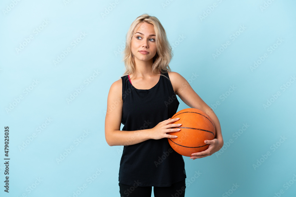 Young Russian woman playing basketball isolated on blue background making doubts gesture looking side