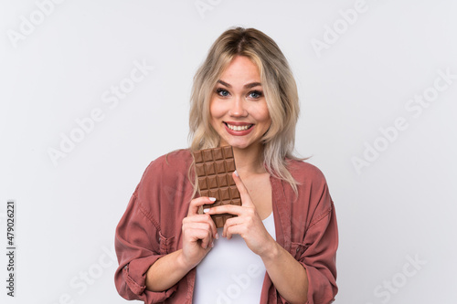 Teenager blonde girl over isolated white background taking a chocolate tablet and happy
