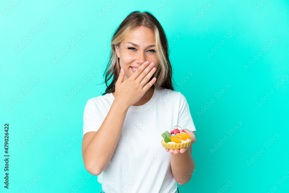 Young Russian woman holding a fruit sweet isolated on blue background happy and smiling covering mouth with hand