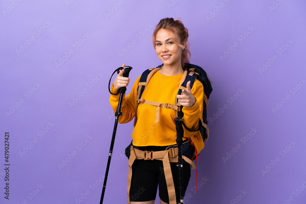 Teenager girl with backpack and trekking poles over isolated purple background pointing to the front and smiling