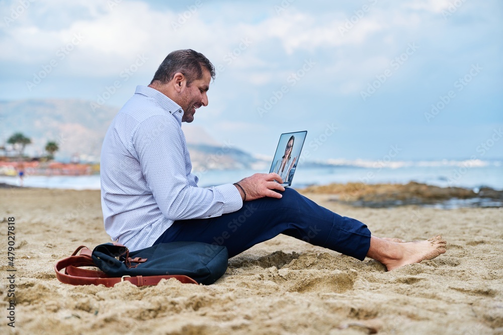 Mature businessman talking with female using video call on laptop sitting on beach