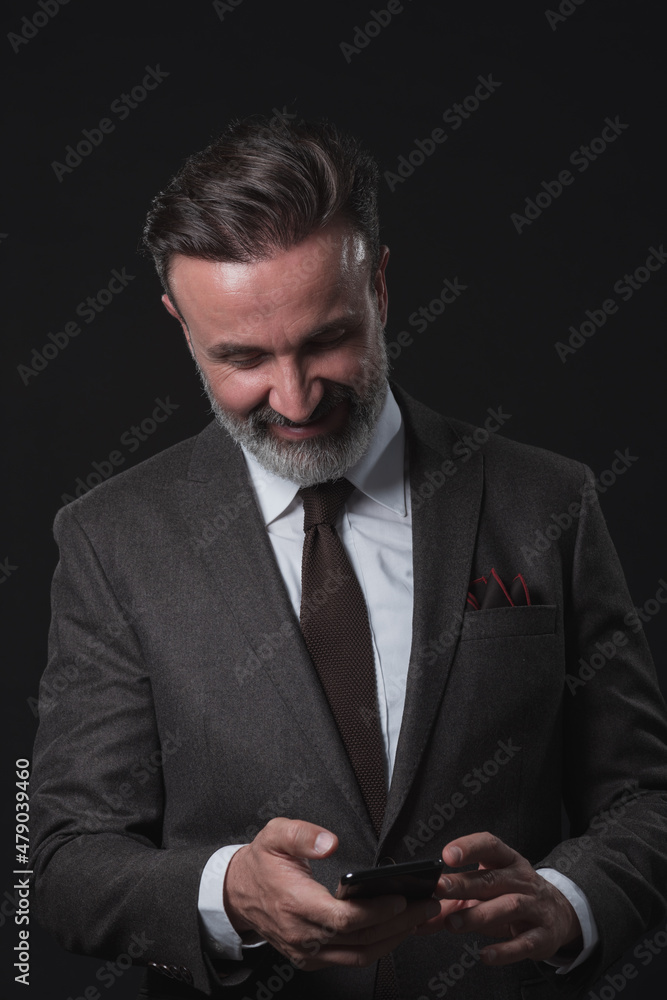 Chat with client. Good looking young man in full suit using his smart phone and smiling while standing against grey background. Chat with client. Good looking young man in full suit using his smart phone and smiling while standing against grey background.
