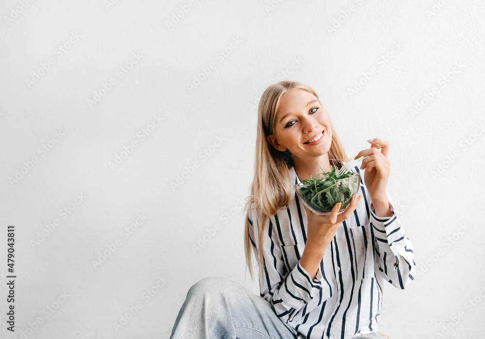 Happy blonde model girl eating fresh salad from a bowl and looking to at camera Happy blonde model girl eating fresh salad from a bowl and looking to at camera