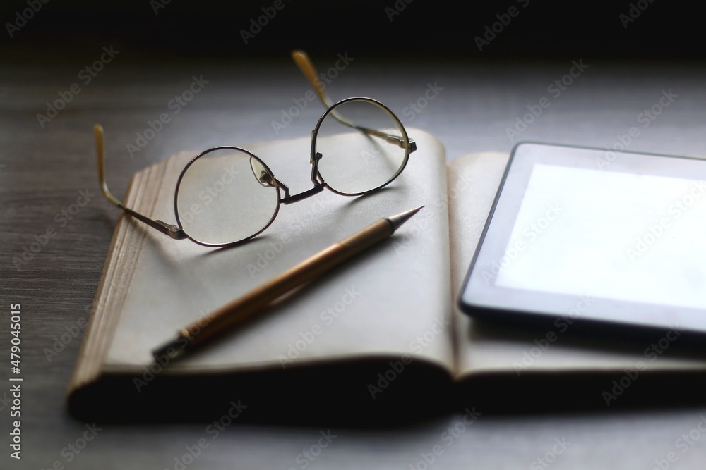 Open book, reading glasses, golden mechanical pencil and e-reader device on the table. Selective focus. Open book, reading glasses, golden mechanical pencil and e-reader device on the table. Selective focus.