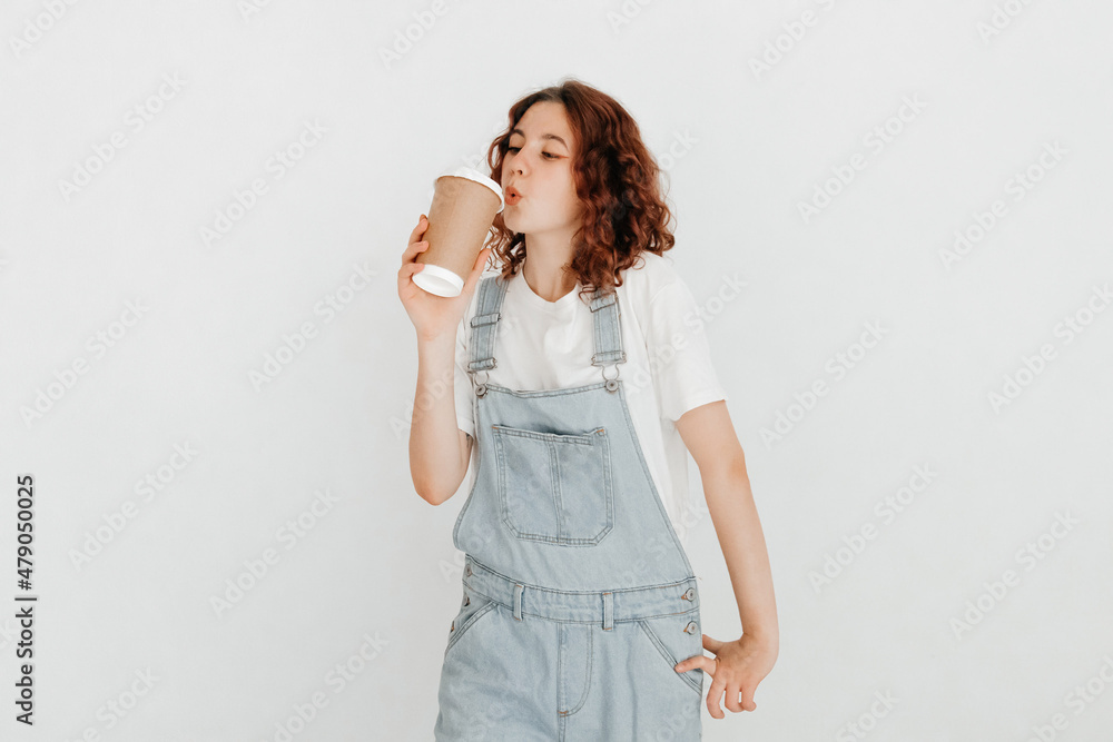 Cheerful redhead girl, wearing denim dungarees and white t-shirt, drinking  hot beverage from a coffee cup, glad to finish studying over white  background Stock-Foto | Adobe Stock