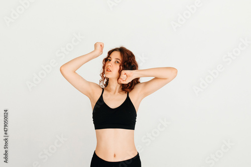 Young redhead yoga instructor stretching her wrists, wearing black top, standing at white background, looking at the camera. Morning exercise © ChesterAlive91