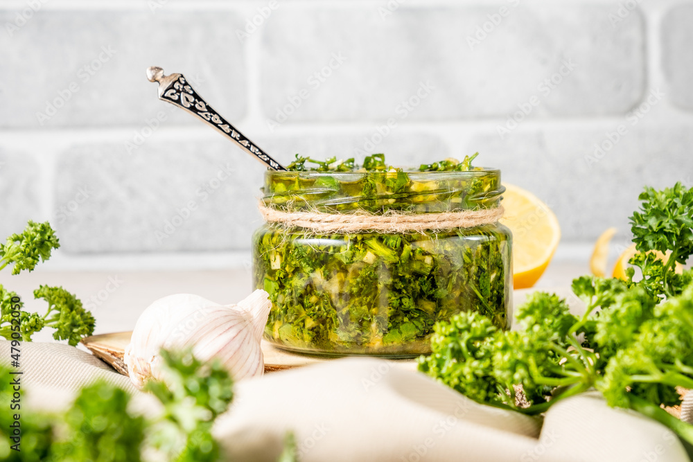 Classic chimichurri sauce based on olive oil, herbs and spices in a glass jar against a gray brick wall. Classic chimichurri sauce based on olive oil, herbs and spices in a glass jar against a gray brick wall.