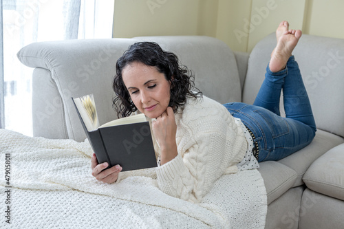 Mujer joven y morena tumbada en le sofá de casa leyendo un libro