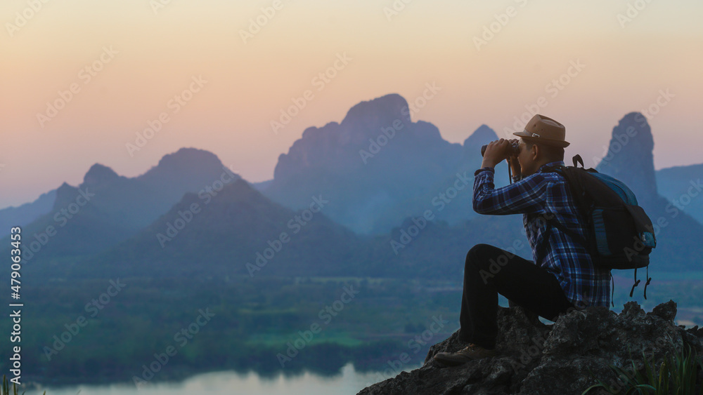 Hiker is sitting and looking sunset on top of mountain with packpack and holding the binoculars.Concept of adventure travel