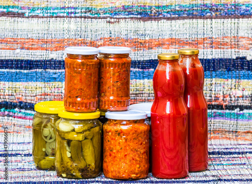 Glass jars with pickled red bell peppers and pickled cucumbers (pickles) isolated. Jars with variety of pickled vegetables, jars with zacusca and bottles with tomatoes sauce. Preserved food concept. photo