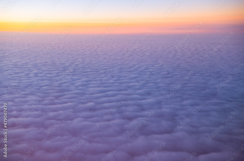 view of sea of clouds at sunset out of airplane window seat window orange sunset with pink and purple fluffy clouds as seen while in flight on airplane our of plane window heavenly horizontal format 