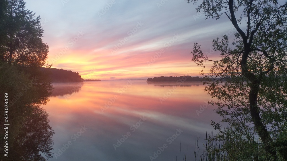 On a spring morning, the sun rises over the lake and illuminates the clouds. The sky and clouds are reflected in the calm water. Reeds grow in the water. There are young leaves on the trees 