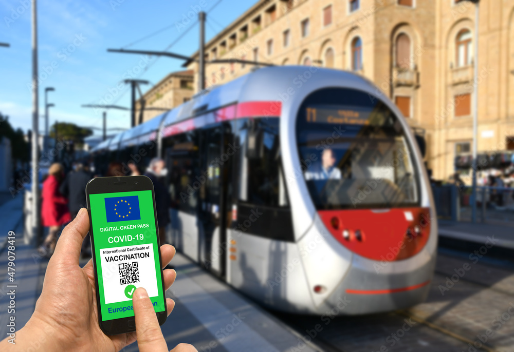 A man at a tram stop is holding smartphone with the European Union digital Green Pass for Covid-19 SARS-CoV-2. Safe travel concept during the Coronavirus pandemic and the Green Pass.