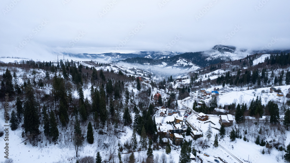 Aerial winter landscape with small village houses between snow covered forest in cold mountains