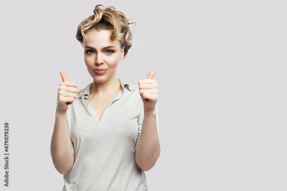 Smiling young woman with thumbs up. Beautiful blonde in a gray shirt. Success and positive. Isolated on a light gray background. Space for text. Smiling young woman with thumbs up. Beautiful blonde in a gray shirt. Success and positive. Isolated on a light gray background. Space for text.