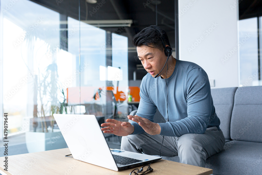 Happy and smiling Asian man in a modern office uses a headset for a video call, a businessman smiles at the interlocutor, and looks at the laptop screen Happy and smiling Asian man in a modern office uses a headset for a video call, a businessman smiles at the interlocutor, and looks at the laptop screen