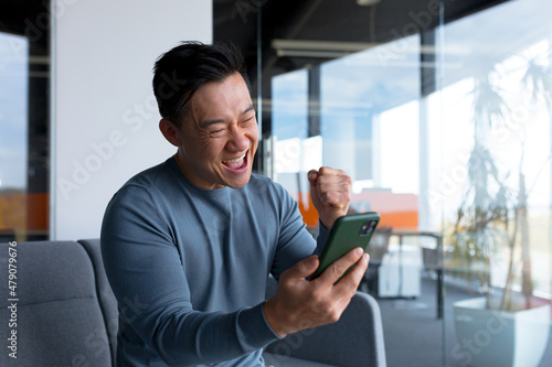 Successful and happy asian businessman man celebrating victory sitting and working in modern office at desk, celebrating victory looking at camera and joyfully shouting holding mobile phone