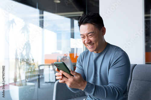Successful and happy asian businessman man celebrating victory sitting and working in modern office at desk, celebrating victory looking at camera and joyfully shouting holding mobile phone