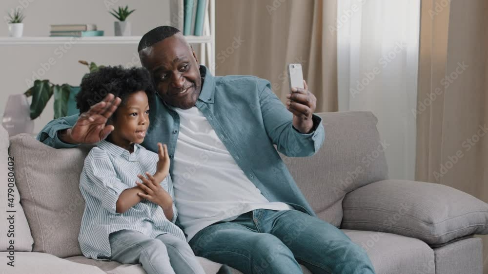 African American family father and daughter sitting in room on couch chatting on webcam mature man with little cute girl shoots video for blog looking at smartphone screen waving hello to phone camera African American family father and daughter sitting in room on couch chatting on webcam mature man with little cute girl shoots video for blog looking at smartphone screen waving hello to phone camera