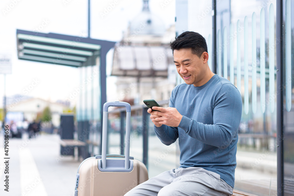 Tourist asian man smiles and looks at the camera sitting at a public transport stop, uses the phone and enjoys a big suitcase Tourist asian man smiles and looks at the camera sitting at a public transport stop, uses the phone and enjoys a big suitcase