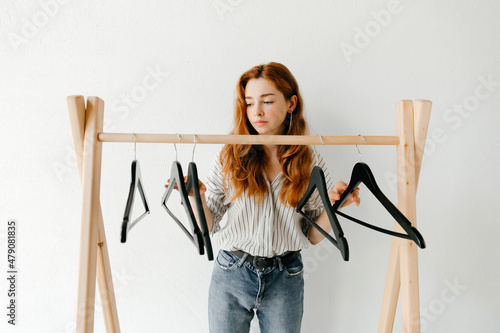 Beautiful disappointed young woman with empty clothes hangers on wooden clothes rack, minimalistic consumption of goods concept.