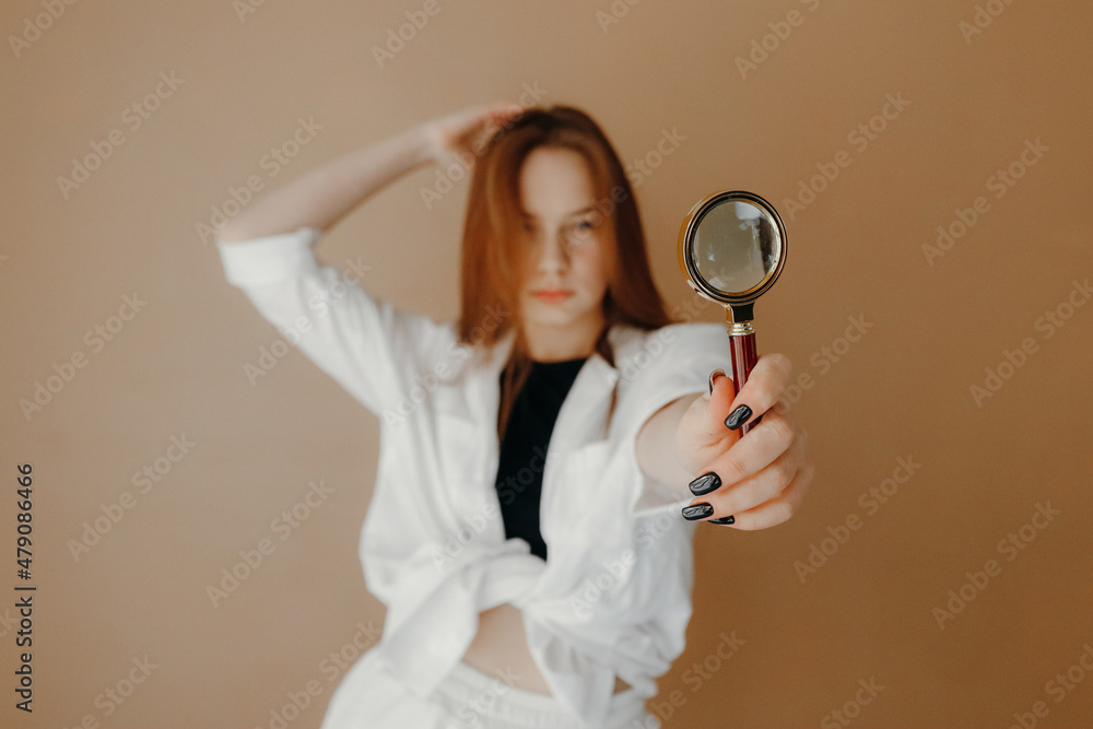 Selective focus, pretty woman in white shirt looking at camera through magnifying glass, isolated over brown background Selective focus, pretty woman in white shirt looking at camera through magnifying glass, isolated over brown background