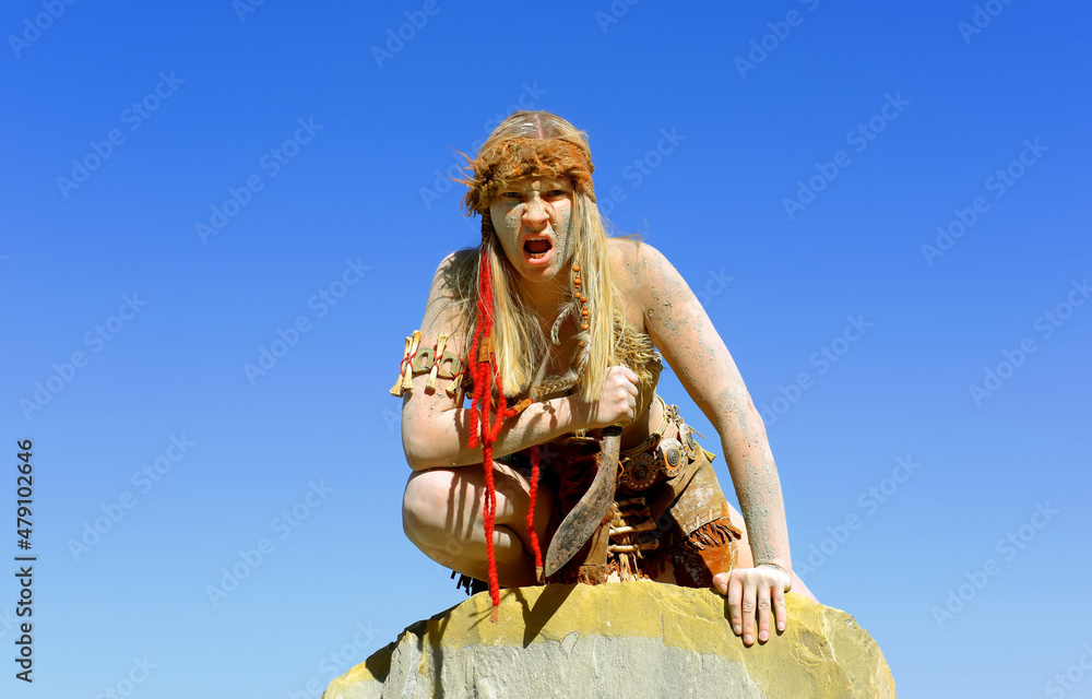 Fototapeta premium A young woman is dressed as Neanderthal warrior. Her body and face is covered with mud and dirt. She is seen posing in a stone quarry. 