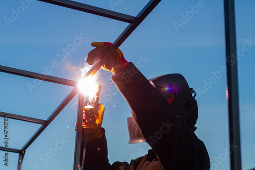 A man in a mask welds the construction. Industrial worker at a welding plant. A strong man - a welder, a welding mask, welds a metal product with arc welding, blue sparks fly to the sides.