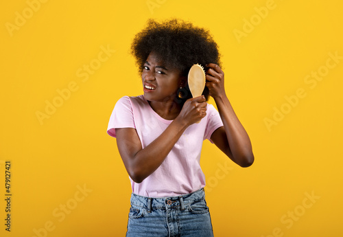 Domestic hairdressing concept. Frustrated african american woman trying to brush her tangled hair, yellow background